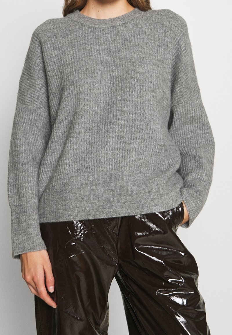 Femme portant un pull en maille côtelée gris et un pantalon en vinyle brun foncé brillant, une main dans la poche, sur un fond uni.