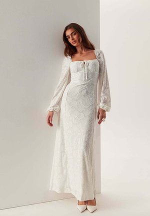 Kvinde iført lang hvid blomstret blondekjole med puffede lange ærmer og firkantet udskæring, stående mod en todelt hvid væg.