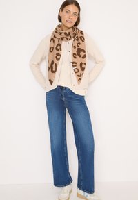 Beige cardigan over en lys sweater, parret med brede blå jeans og et leopardmønstret tørklæde. Hvide sneakers med lyseblå accenter.