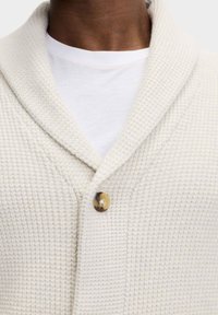Waffelstrick-cremefarbener Cardigan mit Schalkragen, einfachem Rundknopfverschluss und strukturiertem Stoff, über einem weißen Rundhalsausschnitt-Shirt getragen.