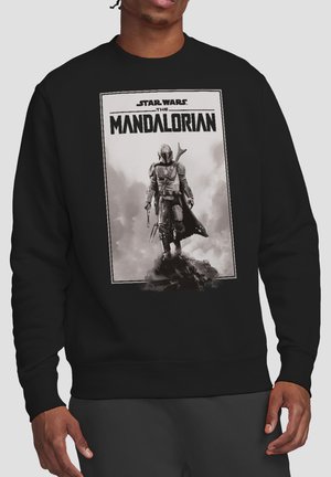 Person mit schwarzem Sweatshirt mit schwarz-weißem Motiv eines Mandalorian-Charakters, der auf einem Felsen steht, unter dem Schriftzug Star Wars The Mandalorian.