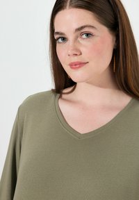 Olijfgroene V-hals top met korte mouwen, gemaakt van zachte stof, met een gladde textuur en subtiele stikdetails langs de halslijn.