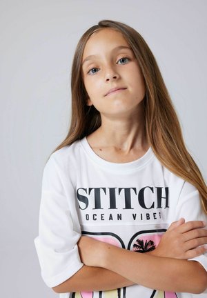 Joven niña con cabello largo castaño claro cruza los brazos, vistiendo una camiseta blanca con la inscripción "STITCH OCEAN VIBES" y gráficos de palmeras.