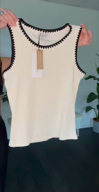 Sleeveless white top with black scalloped trim on neckline and armholes held up by person indoors, tags attached, with plants in the background.