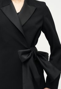 Blazer noir sur mesure avec de larges revers en satin et un grand nœud satinés noué à la taille, porté par-dessus un haut noir.