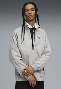 Chaqueta gris claro con un diseño de media cremallera, panel frontal texturizado, puños elásticos y logo negro. Tejido suave con un corte relajado.