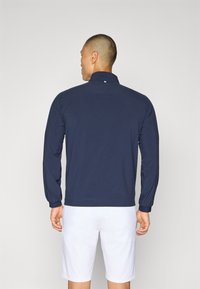 Long-sleeve navy sweatshirt with a textured pattern, high collar, and elastic cuffs, paired with white shorts. No visible logos or graphics.