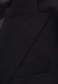 Detail van een zwarte colbert met gestructureerde stof, geknopt revers en een enkele borstzak met stikaccenten.