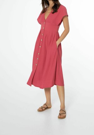 Femme portant une robe rose boutonnée au genou avec des manches courtes et des sandales, debout avec une main dans la poche sur un fond blanc.