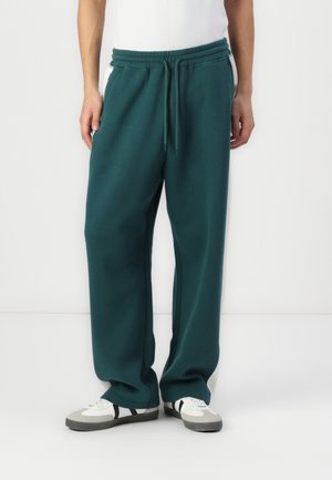 Personne portant un pantalon de jogging ample vert foncé et des baskets blanches avec des rayures noires, debout devant un fond uni.