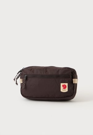 Riñonera compacta de color marrón oscuro con bolsillo frontal con cremallera y parche de logo beige con la cabeza de un zorro rojo sobre fondo claro.