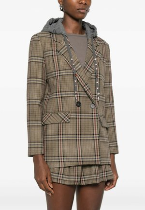 Donna che indossa un blazer marrone a quadri con doppi bottoni, gonna coordinata e una felpa grigia con cappuccio sotto.