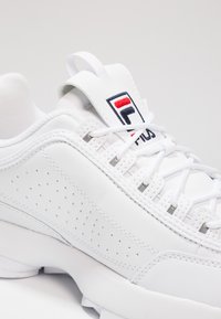 Sneaker blanche avec tige en cuir perforé, lacets, languette rembourrée et un patch logo en bleu, rouge et noir. Semelle en caoutchouc avec dessin antidérapant.