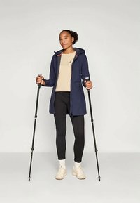 Mornarsko vodoodporno jopico s kapuco, kontrastnimi koralnimi akcenti, sprednjimi žepi, oblečeno čez bež majico in črne tajice, z pohodnimi palicami.