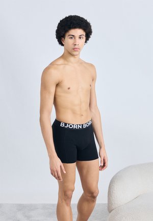 Mustad bokserid valge "BJORN BORG" vöökohaga, tiheda lõikega, sileda tekstuuriga, kandmas afro-soenguga inimene.