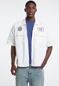 Giacca bianca a maniche corte con due tasche frontali e toppe ricamate; indossata sopra una t-shirt blu e abbinata a jeans azzurri chiari.