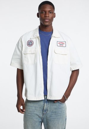 Weißer Kurzarmjacke mit zwei Fronttaschen und gestickten Patches; getragen über einem blauen T-Shirt und kombiniert mit hellblauen Jeans.