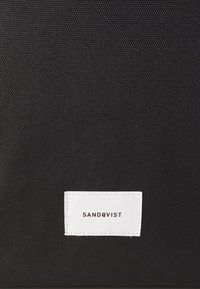 Czarna torba z materiału o teksturowanej powierzchni, z białą prostokątną metką z napisem "SANDQVIST". Prosty i funkcjonalny design.