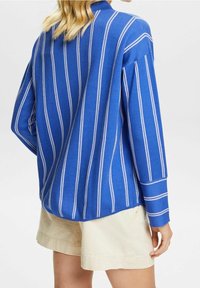 Camisa de manga larga azul con rayas verticales blancas, de tejido suave, corte relajado y con cuello. Combinada con pantalones cortos de color claro.