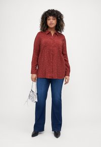 Chemise rouge à boutons avec des pois blancs, manches longues, poches poitrine, assortie à un jean évasé bleu et un sac à main argenté.