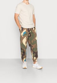 Beige T-shirt met korte mouwen en zak, gecombineerd met joggers in camouflagepatroon, met groene, bruine en blauwe accenten. Witte sneakers maken de look compleet.
