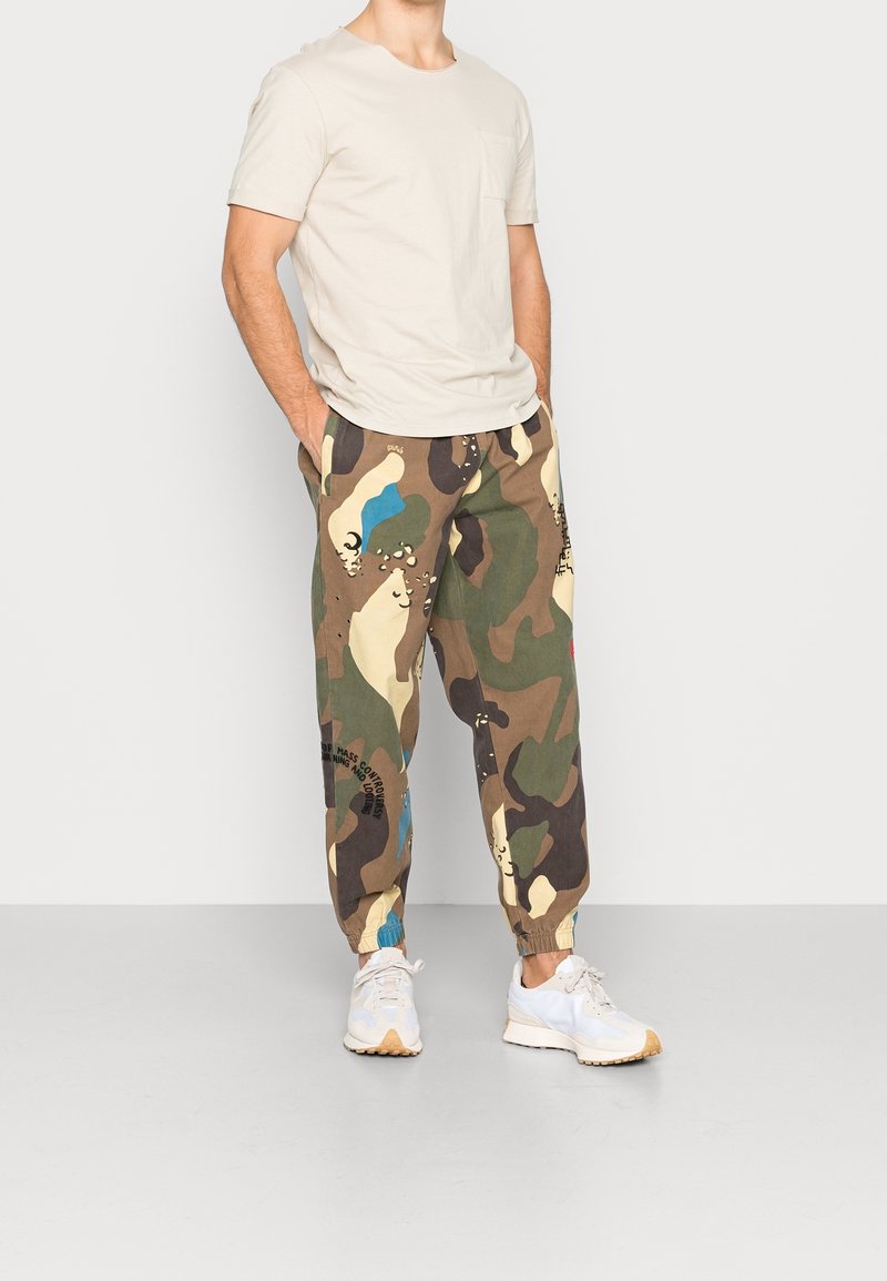 Beige T-shirt met korte mouwen en zak, gecombineerd met joggers in camouflagepatroon, met groene, bruine en blauwe accenten. Witte sneakers maken de look compleet.