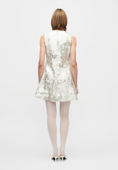 Rochie scurtă argintie cu siluetă evazată, model floral texturat, fără mâneci și închidere cu fermoar la spate; asortată cu colanți albi și pantofi cu toc.