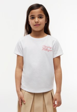 Młoda dziewczyna z długimi ciemnymi włosami, ubrana w białą koszulkę Tommy Hilfiger i beżową plisowaną spódnicę, stoi na jednolitym białym tle.