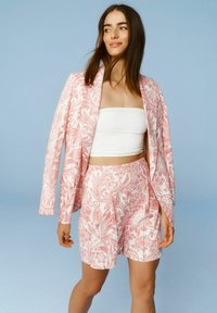Compleu de sacou și pantaloni scurți cu imprimeu floral în nuanțe de roz deschis. Sacoul are o croială lejeră și mâneci lungi, iar pantalonii scurți sunt adaptați cu pliuri. Sub sacou se află un top alb scurt.