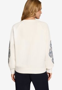 Femme vue de dos portant un sweat-shirt blanc avec des motifs de perles en forme de cœur bleu sur les deux manches, associée à un pantalon foncé.