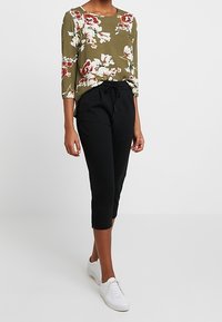 Haut vert à imprimé floral avec des manches trois-quarts, associé à un pantalon noir court et des baskets blanches, doté d'une taille à cordon.