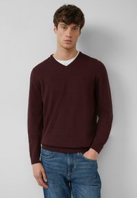 Burgunderfarbener V-Ausschnitt-Pullover aus feinem Strickstoff, mit gerippten Bündchen und Saum; über einem weißen T-Shirt getragen; kombiniert mit blauen Jeans.