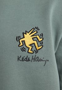Green cotton fabric features a yellow embroidered figure with outstretched limbs, accompanied by the text "Keith Haring™" below.