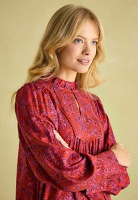 Red floral patterned blouse with a high neckline, gathered bodice, and long puffed sleeves. Features a keyhole opening at the front.