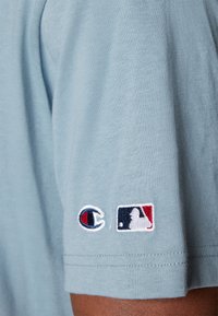 Gaiši zils īsām piedurknēm T-krekls, kas izgatavots no mīkstas auduma, ar izšūtiem logo: Champion logo un MLB simbols kreisajā piedurknē.