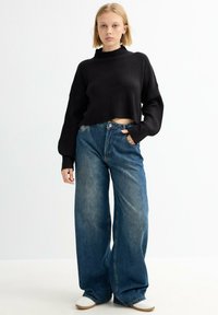 Mujer rubia vestida con un suéter negro de canalé, jeans de pierna ancha azul y zapatillas blancas, de pie frente a un fondo blanco liso.