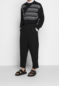 Svart tröja med vitt textmönster, lösa svarta byxor och svarta sandaler med tjocka remmar och texturerade sula.