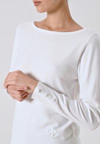 Maglione bianco a maniche lunghe realizzato in tessuto morbido, con un ampio scollo e dettagli con bottoni sui polsini, e un logo sottile vicino all'orlo.