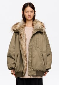 Mujer con una chaqueta bomber de gran tamaño en color verde oliva, con un gran cuello de piel y bolsillos con cremallera, combinada con un suéter claro y pantalones oscuros.