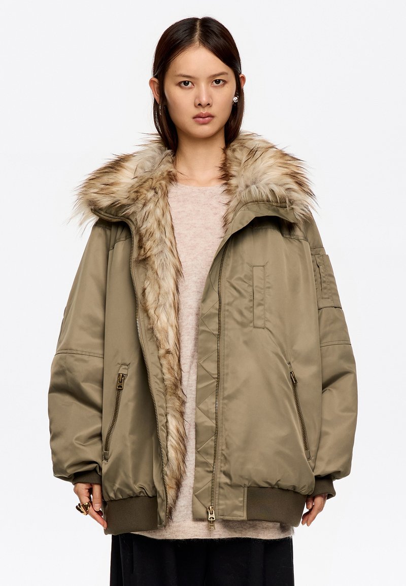 Mujer con una chaqueta bomber de gran tamaño en color verde oliva, con un gran cuello de piel y bolsillos con cremallera, combinada con un suéter claro y pantalones oscuros.