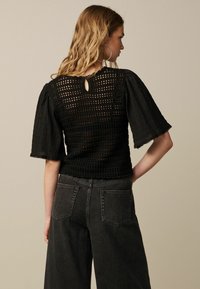 Top all'uncinetto nero con maniche a sbuffo, caratterizzato da un'apertura a chiave sul retro. Abbinato a jeans grigi scuri a vita alta. Tessuto testurizzato in tutto il capo.