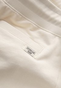 Gros plan d’un tissu beige avec une ceinture côtelée et une petite étiquette cousue portant la mention « You only limit your mind – OMBRE depuis 1985. »
