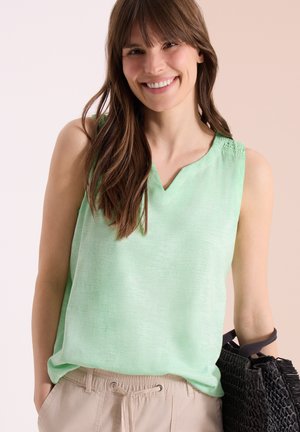 Femme souriante, portant un haut sans manches vert clair et un pantalon beige, tenant un sac tissé noir avec une main dans la poche.