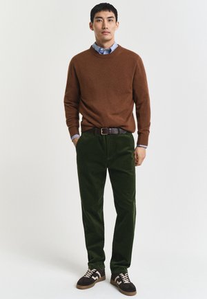 Mann steht und trägt einen braunen Pullover über einem blauen Hemd mit Kragen, dunkle grüne Hose, einen schwarzen Gürtel und braune Sneakers mit weißen Schnürsenkeln.