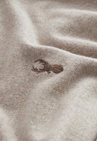 Beige strikket stof med en blød tekstur; pryder en lille broderet hummerdesign i brun på overfladen.