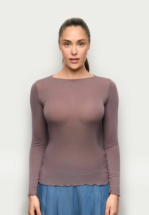 Kobieta o ciemnych włosach związanych w kucyk, ubrana w dopasowaną, fioletoworóżową bluzkę z długim rękawem i niebieską spódnicę, stojąca przodem na neutralnym, jasnym tle.