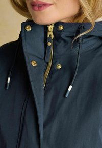 Námornícky modrá bunda so zlatým zipsom, dvoma zlatými cvokmi, kuklou s nastaviteľnými šnúrkami a hladkou, matnou textúrou.