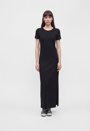 Une femme porte une longue robe noire à manches courtes avec un col rond et des baskets noires et blanches sur un fond clair uni.
