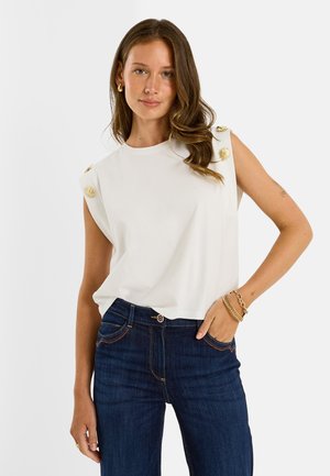 Mujer con cabello largo y ondulado lleva una camiseta blanca sin mangas con botones dorados en los hombros y vaqueros azul oscuro, posando con la mano en el bolsillo.