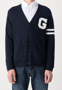 Marinblå stickad cardigan med V-hals, som har en vit 'G' logotyp och ränder på ärmen, två fickor och svarta knappar.